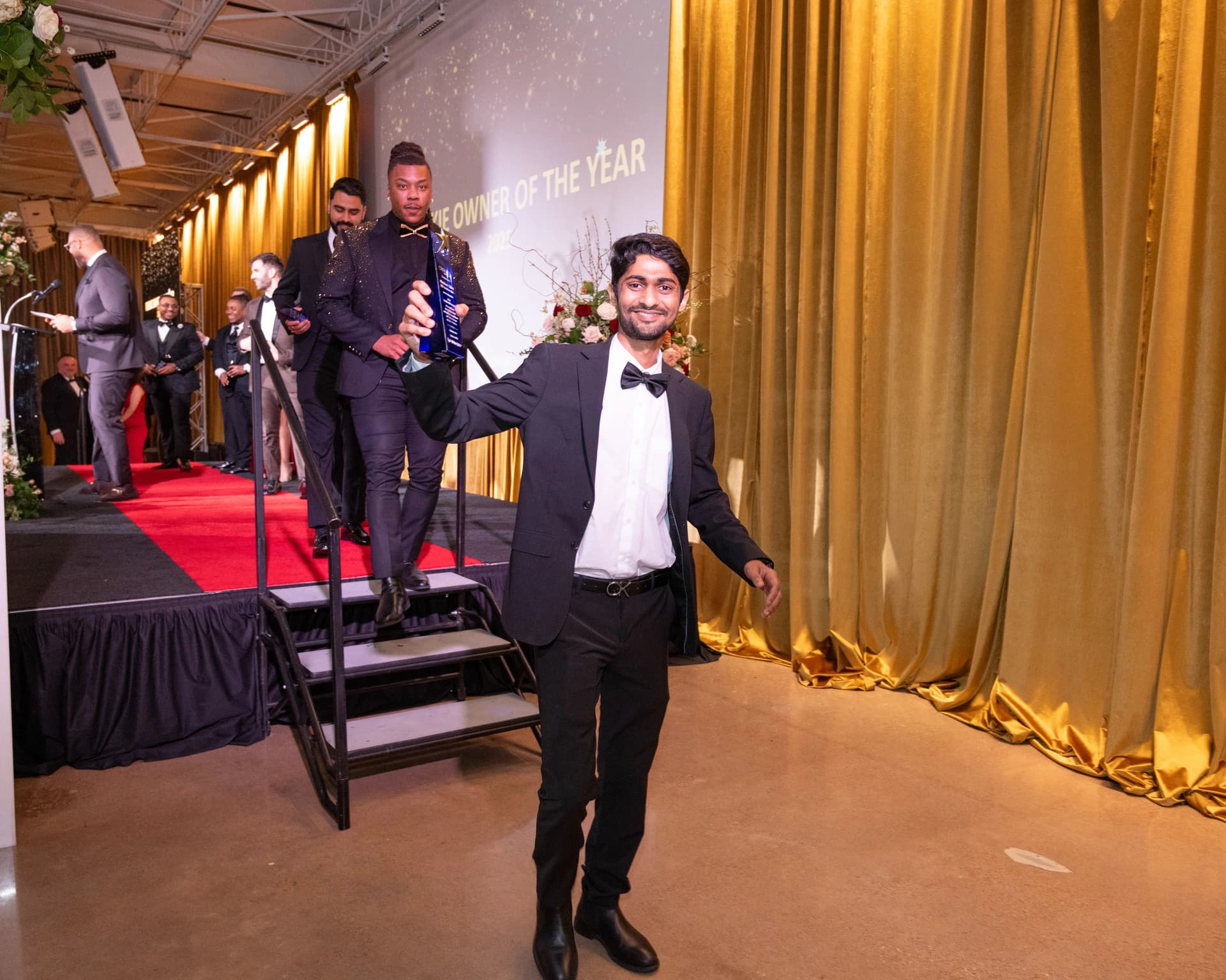 A smiling award recipient holding up his trophy beside the stage at an awards event.