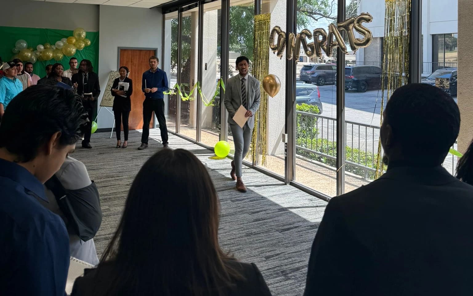 Colleagues gathered around a man holding a document during an office celebration, with gold congratulations balloons behind him.