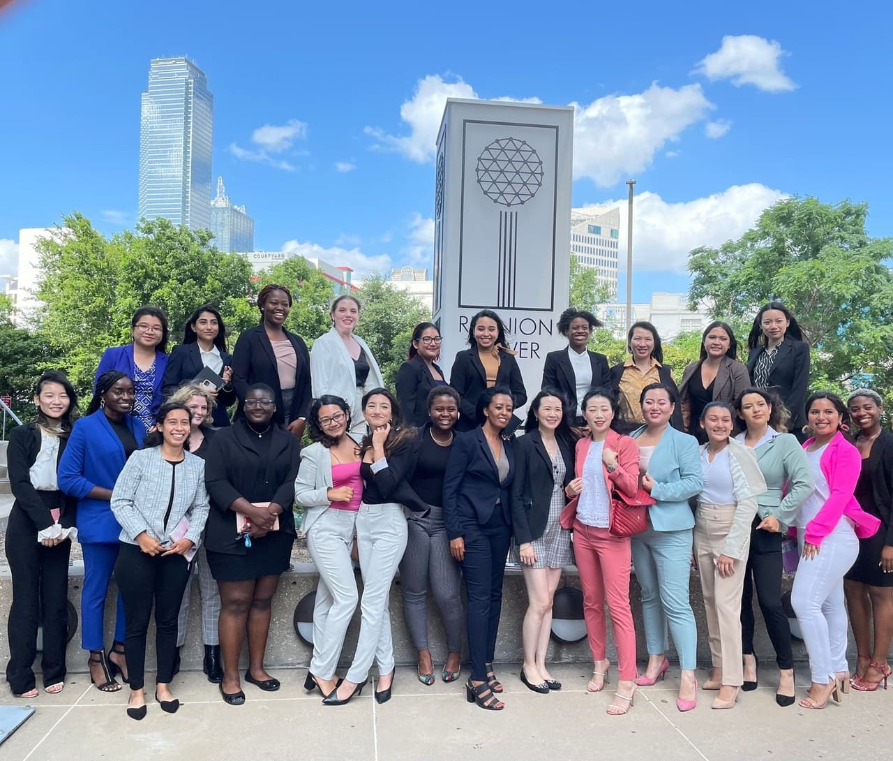 A large team in business dress gathered outdoors in front of a stone sign with a city skyline behind them.