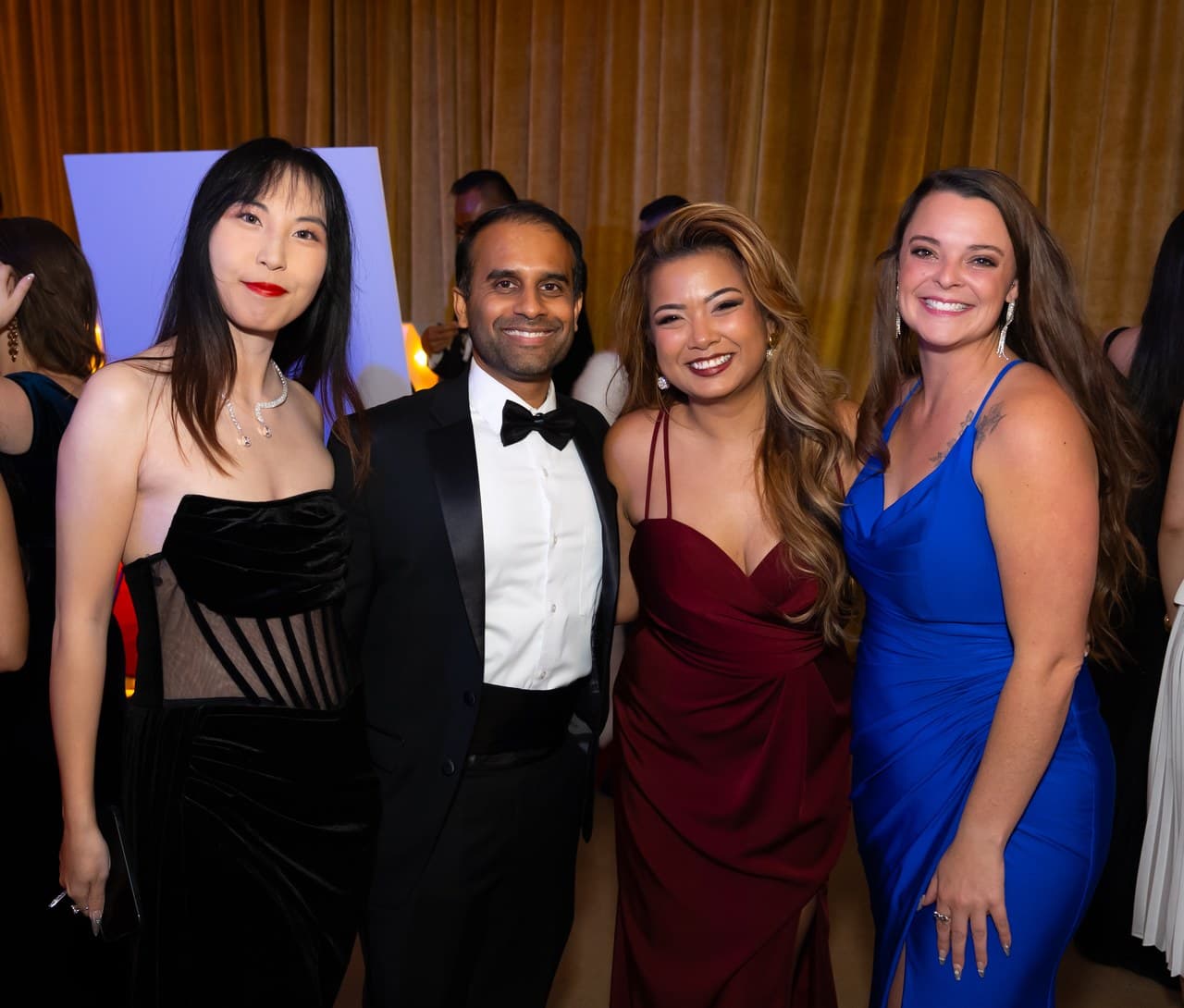 Four colleagues in evening wear smiling together at a formal event in front of gold curtains.