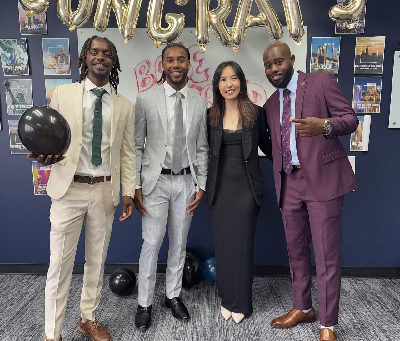 Four team members in suits posing under gold balloon letters that spell congrats.
