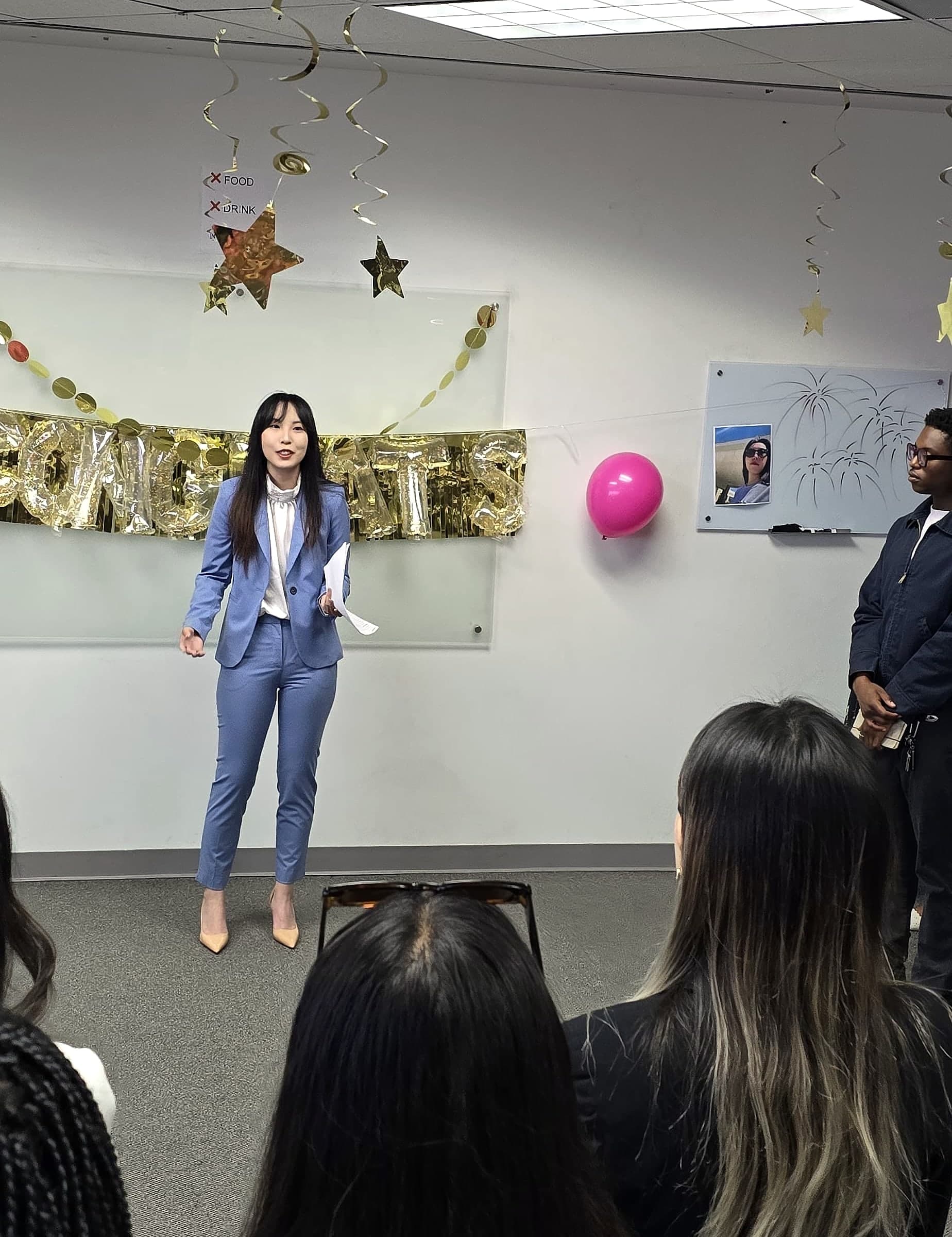 Xichen Mao in a light blue suit speaking to her team in an office decorated with a gold Congrats banner.
