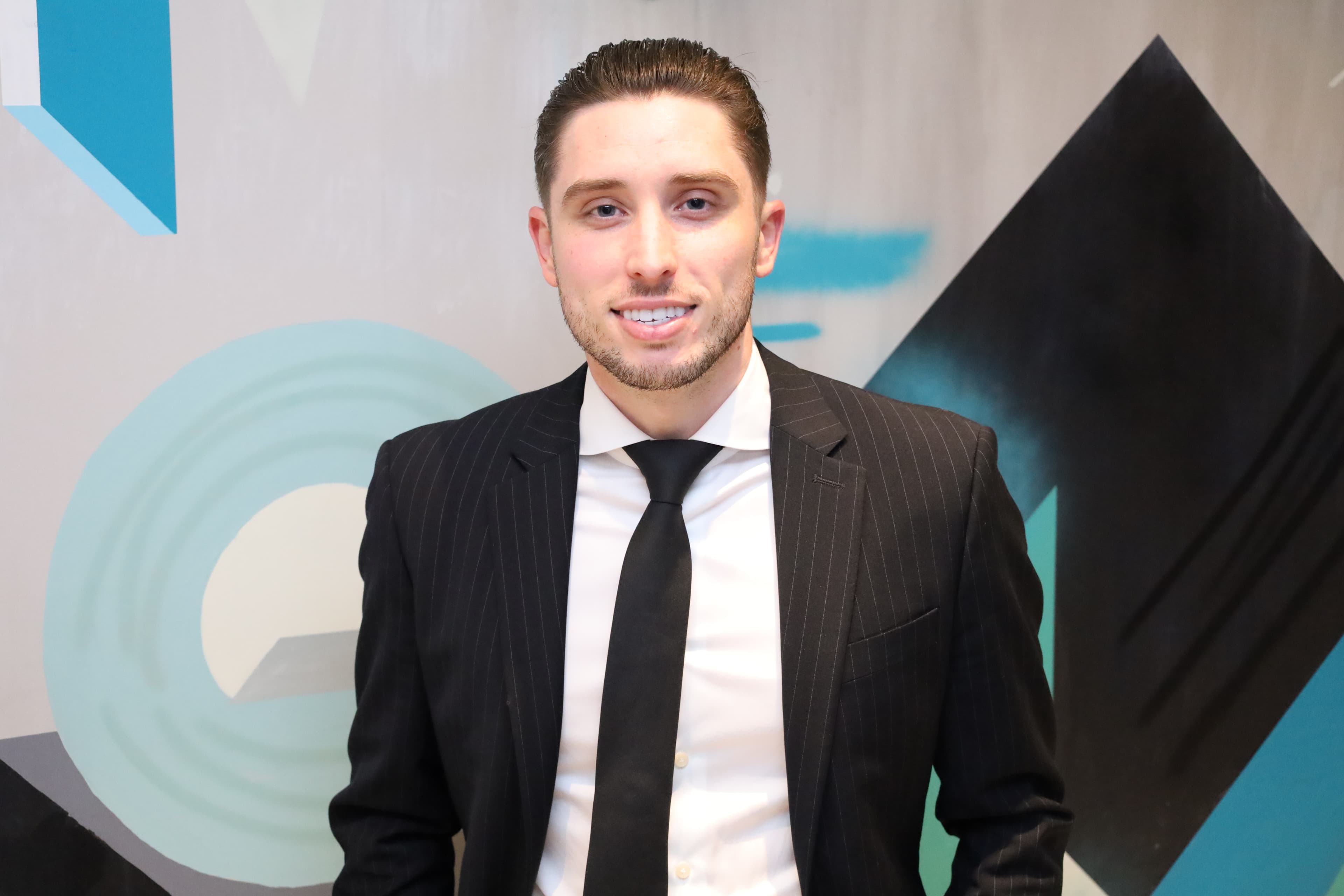 Portrait of Zach Pyron in a dark suit against a blue and charcoal mural