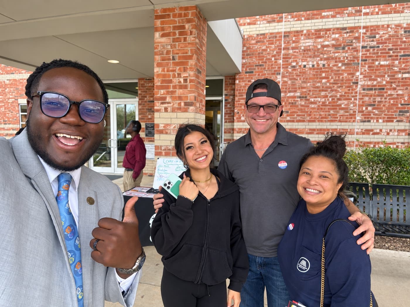 A fundraiser and three supporters outside a brick civic building, one giving a thumbs up