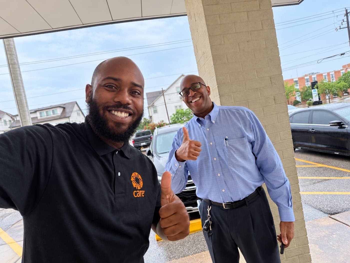 A fundraiser and a supporter giving thumbs up under a covered entrance on a rainy day