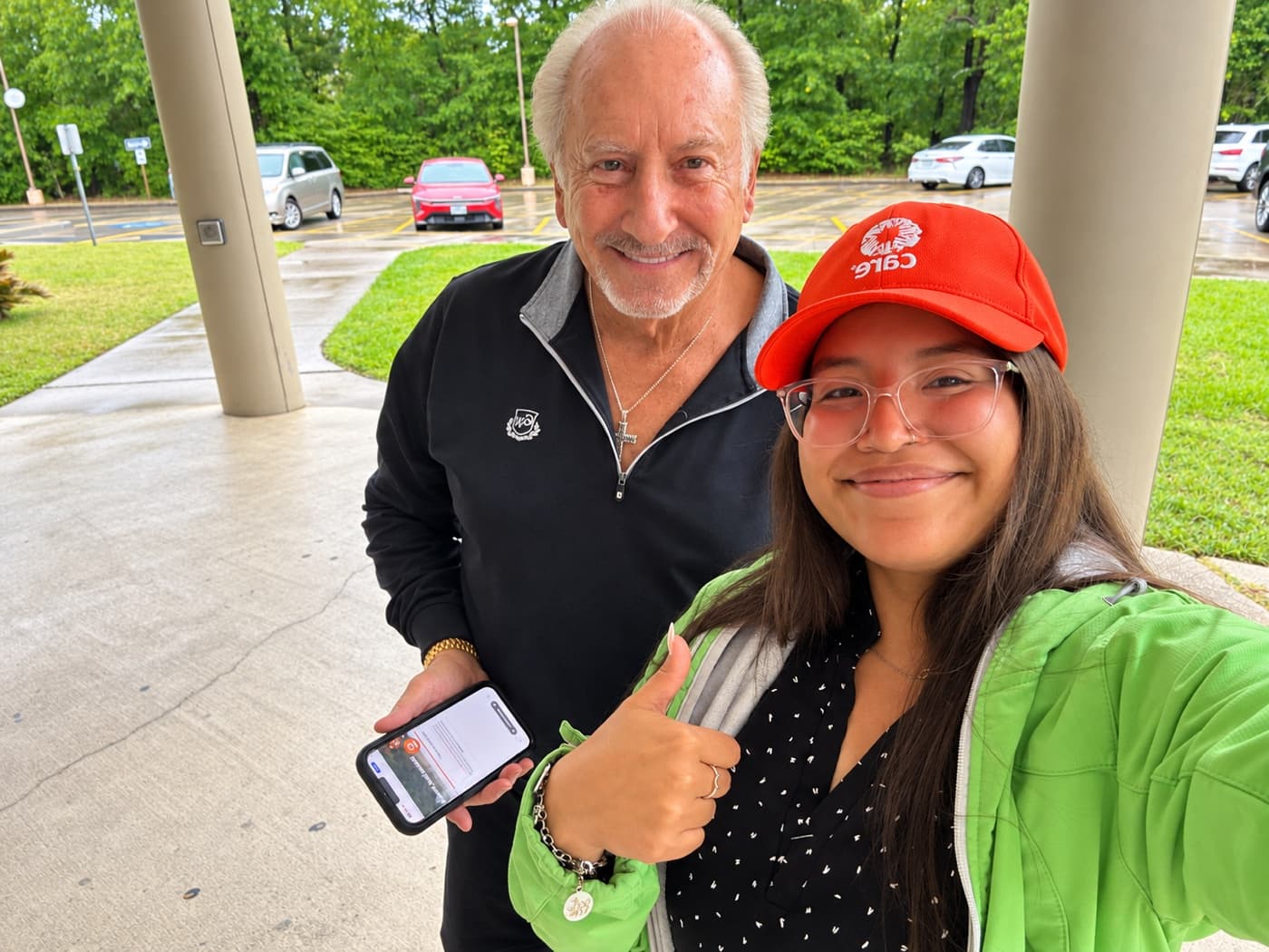 A supporter holding a phone beside a fundraiser giving a thumbs up under a portico