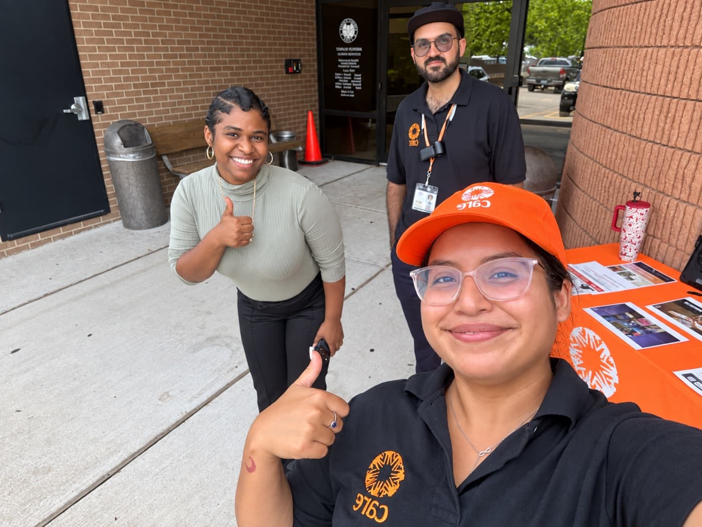 Two fundraisers and a supporter giving thumbs up beside a sign-up table outside a brick building